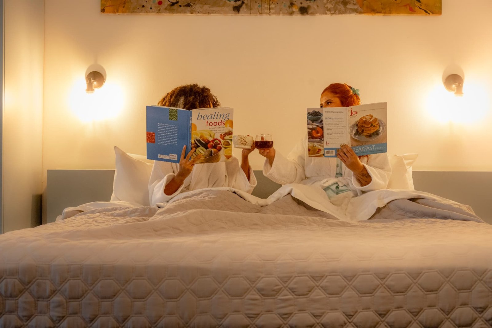 Two women in robes reading and toasting on the bed of a room at Tántalo